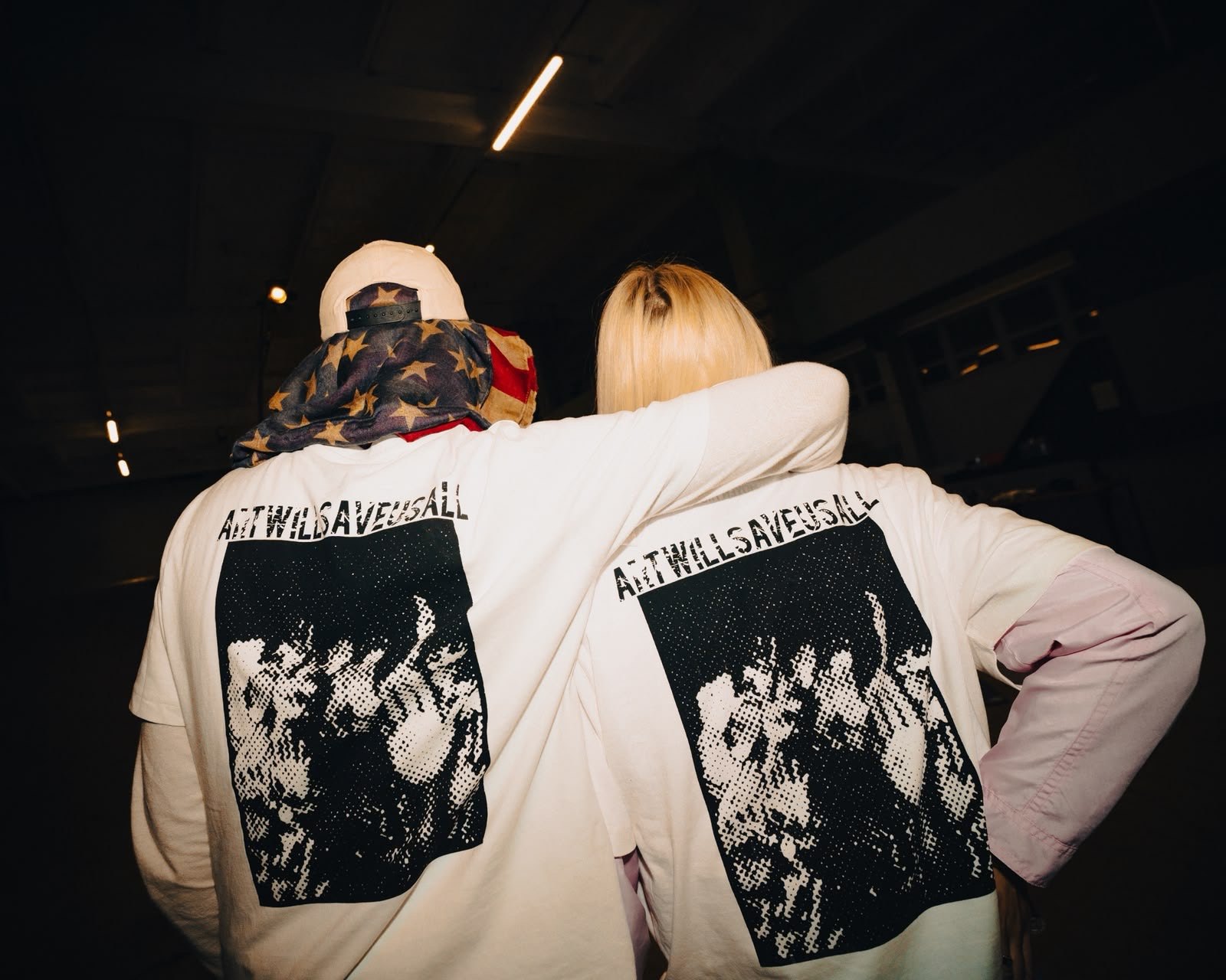 Two people from behind at night wearing the ART WILL TEE, ART WILL SAVE US ALL printed on the back